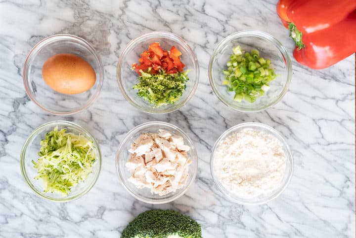 Broccoli stalk, bowl of grated zucchini, bowl of chopped bell pepper and grated broccoli, grater with broccoli, part of a red bell pepper