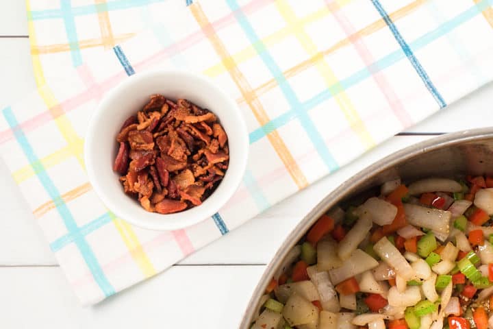 Bowl of cooked bacon under a pastel checkered kitchen towel next to cooked vegetables in a pan