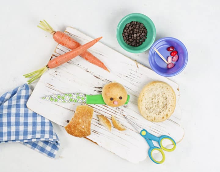 White wooden board with cut cars, cut up bread bowl, bunny face bread bowl, carrots, kitchen towel, scissors, bowl of chocolate chips, bowl of jelly beans