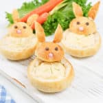 Three cute bunny bread bowls with chowder inside on a white wooden board. Carrots and lettuce as decor in the background.