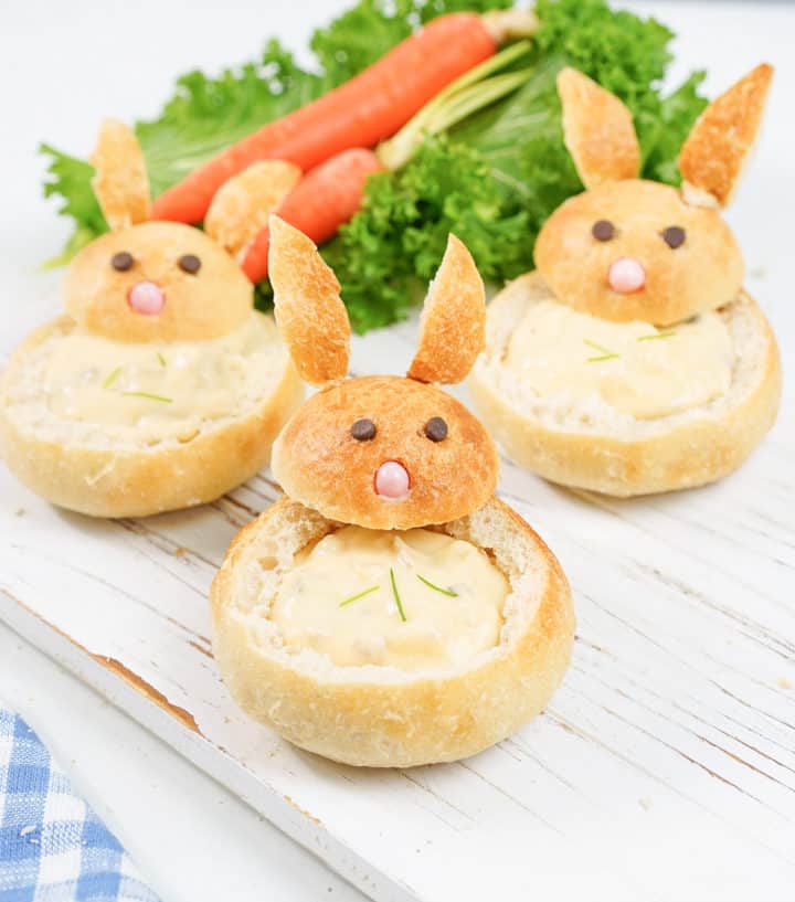 Three cute bunny bread bowls with chowder inside on a white wooden board. Carrots and lettuce as decor in the background.