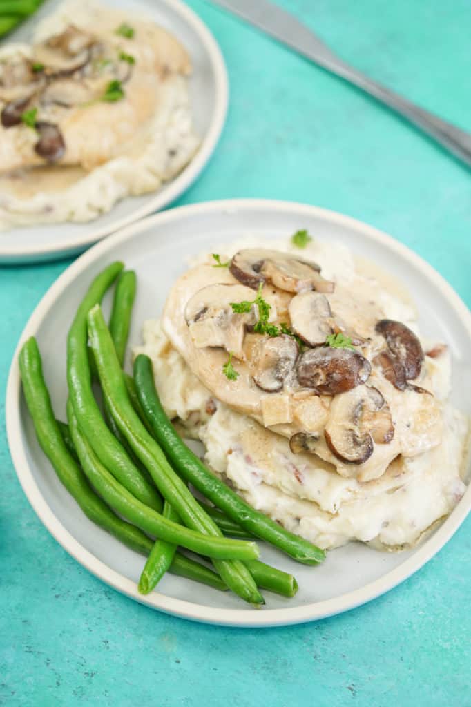 Plate of green beans, creamy mushroom sauce over chicken breast on a bed of mashed potatoes on a teal background