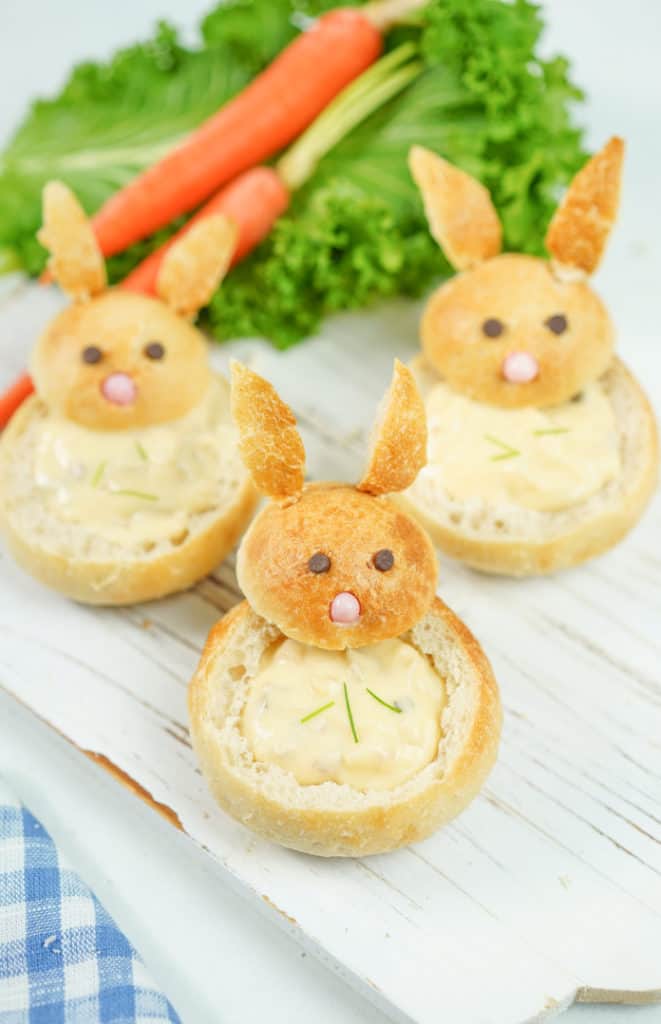 Three carved out bread bowls shaped like bunnies with carrots in the background on white wooden board