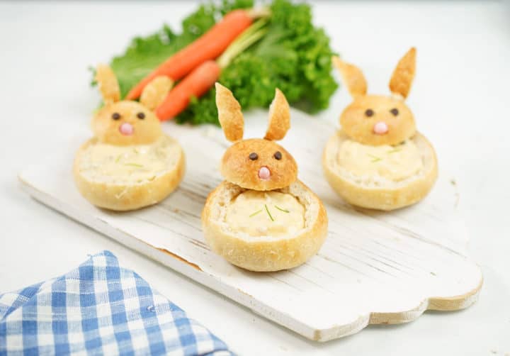 three bunny bread bowls filled with chowder on a wooden board. lettuce and carrots in the background for decor and gingham blue napkin on the left corner