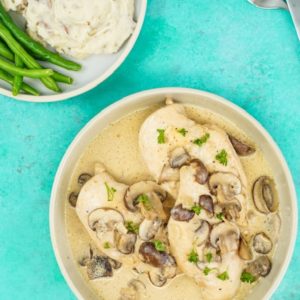 Bowl of chicken with creamy mushroom next to mash potatoes and green beans on a teal background