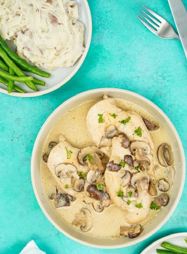 Bowl of chicken with creamy mushroom next to mash potatoes and green beans on a teal background