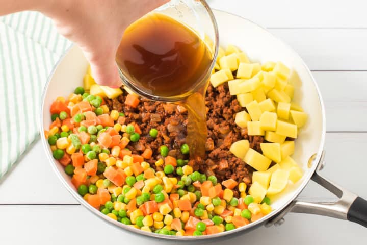 Hand with stock in a glass bowl pouring into a pan of mixed vegetables, ground beef and potatoes 