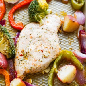 Close up of seasoned chicken next to broccoli, onions, red pepper and potatoes on a sheet pan