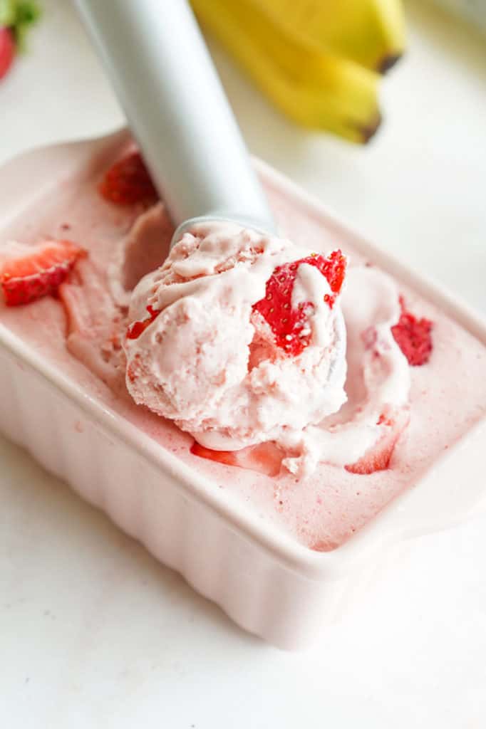 Strawberry ice cream with strawberry chunks in a metal scoper in a small rectangular pink pan