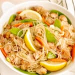 plate of filipino noodles with snow peas, chicken, lemon slices, sliced carrots, sliced cabbage in a bowl