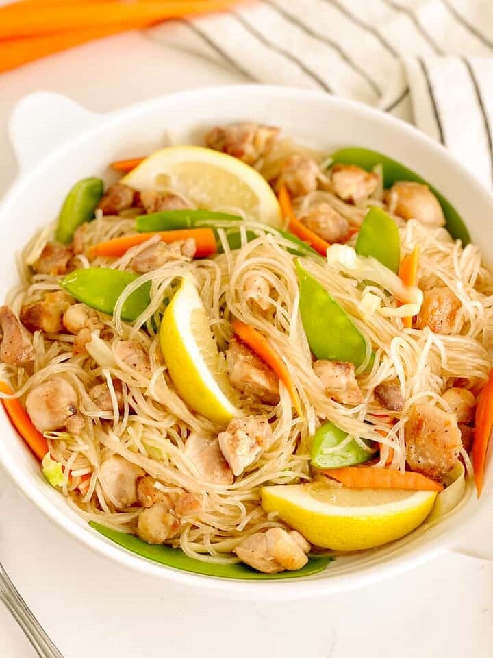 plate of filipino noodles with snow peas, chicken, lemon slices, sliced carrots, sliced cabbage in a bowl