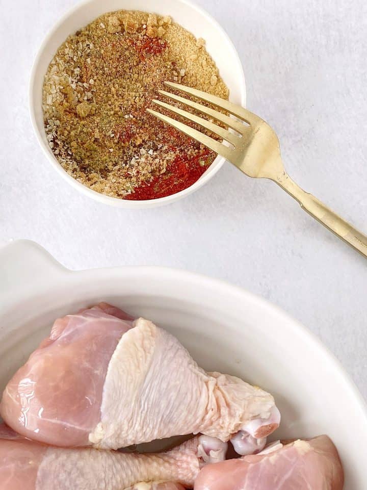 bowl of seasonings with a fork on top next to raw chicken legs in a bowl 
