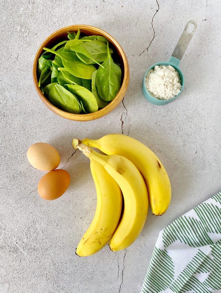 ingredients on a gray surface with bananas and spinach and flour and eggs
