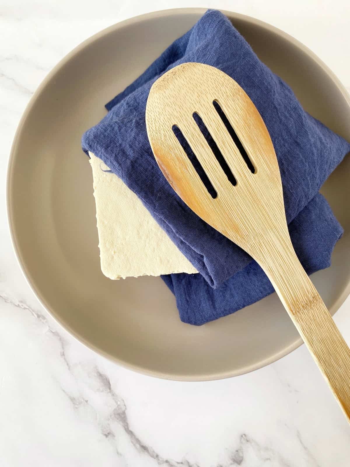 wooden spatula on top of a towel and under is a block of tofu 