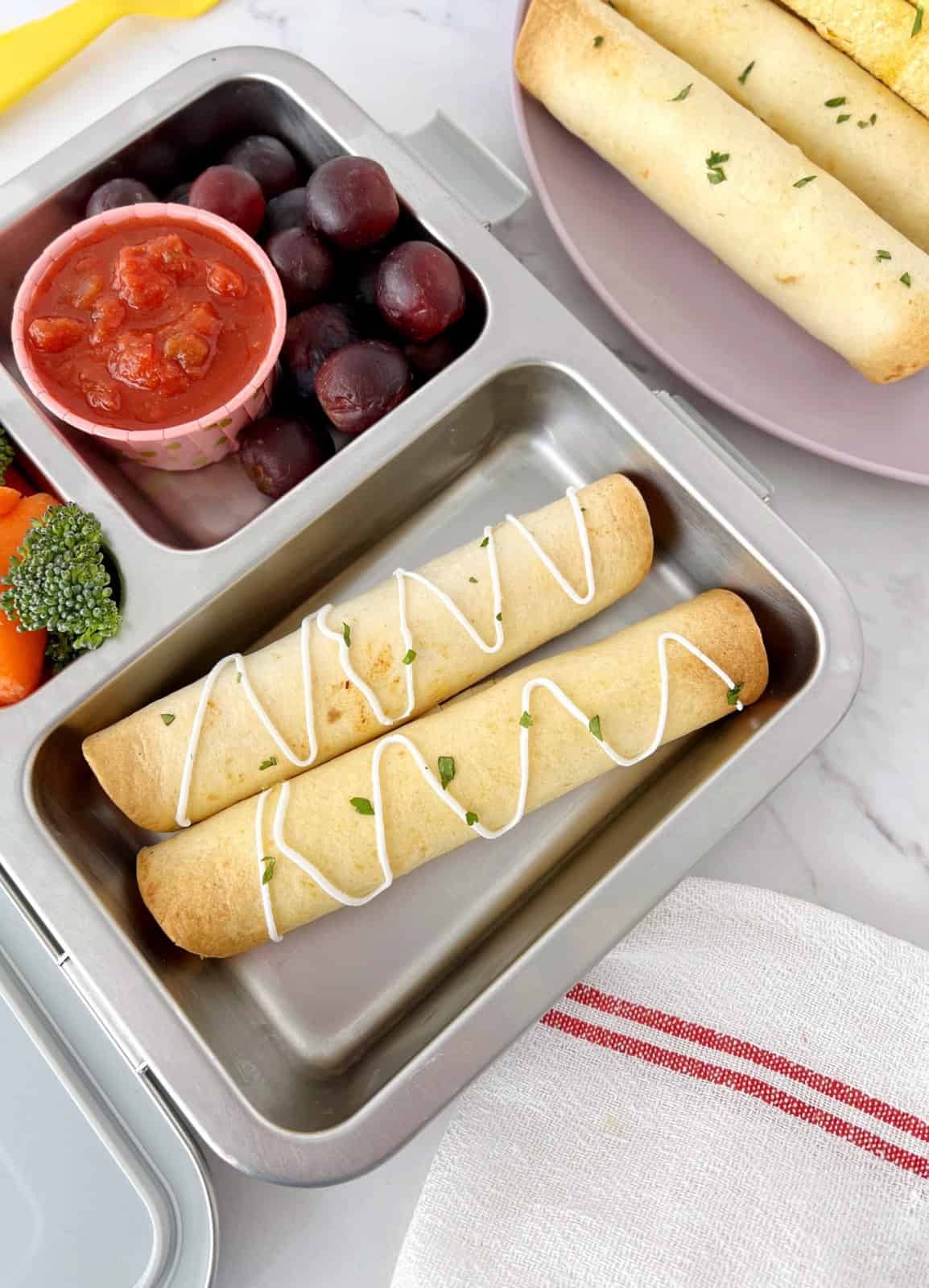 taquitos in a lunch box next to grapes 